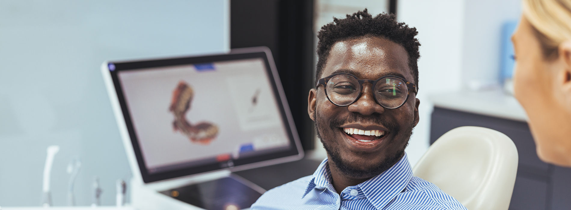 The image shows a man with glasses smiling broadly at the camera while seated in front of a computer screen displaying a graphical representation of a human face, with another person partially visible behind him, suggesting an office environment.