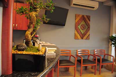 This is a photograph of an interior space, likely a restaurant or cafe, featuring a counter with stools, a potted plant, framed artwork on the wall, a television screen displaying sports content, and a window that allows natural light into the room.