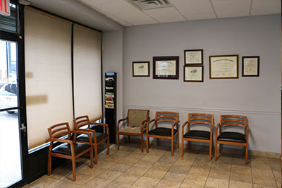 The image shows an interior space with a waiting area featuring chairs, a window with blinds partially open, and framed certificates on the wall behind the chairs.