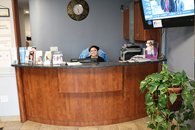 The image shows a reception area with a desk, a chair, a potted plant, and a person sitting at the desk.