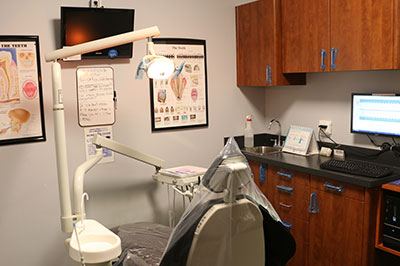 The image shows an interior view of a dental office with a chair, dental equipment, and posters explaining dental services.