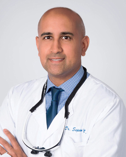The image shows a man wearing a white lab coat with name tags on his chest, standing against a plain background. He has a bald head, a beard, and is smiling at the camera.